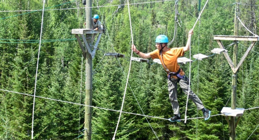 A person wearing safety gear and secured by ropes balances on an obstacle on a high ropes course. 
